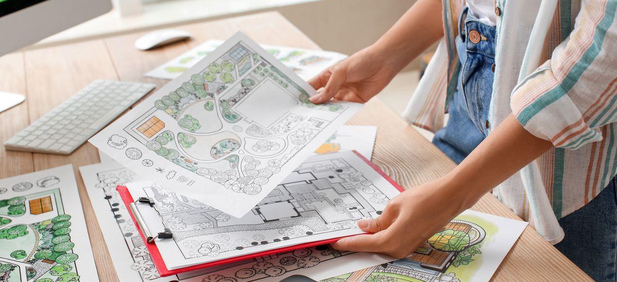 Fachkraft im Gartenbau Walldorf haelt detaillierte Entwurfsplaene fuer die Gestaltung von Aussenanlagen am Schreibtisch in der Planungsphase