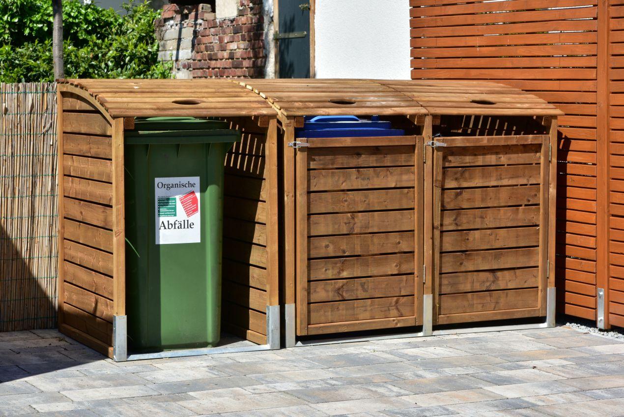 Mülleinhausung aus Holz mit drei Mülltonnen auf einem gepflasterten Bereich, harmonisch integriert in einen natürlichen Gartenzaun